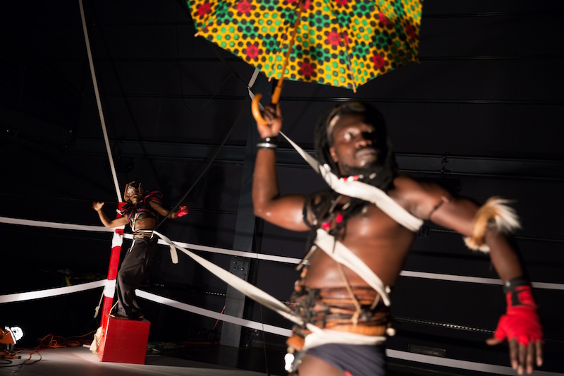 A close up of a shirtless black in a box ring. Harnesses and ropes entangle his torso.